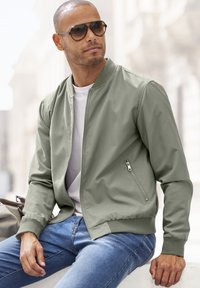 Khaki Bomberjacke mit einem Frontreißverschluss, gerippten Bündchen und einer seitlichen Reißverschlusstasche. Getragen über einem weißen T-Shirt und kombiniert mit blauen Jeans.