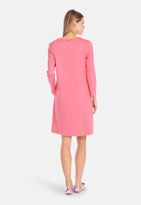 Rosa, jurk tot de knie met lange mouwen, ronde halslijn en een rits aan de achterkant. Glad textuur; gedragen met lichtroze schoenen met strikaccenten.