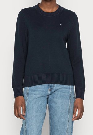 Person, die einen marineblauen Pullover mit langen Ärmeln, gerippten Bündchen und Saum trägt, kombiniert mit hellblauen, hoch taillierten Jeans.