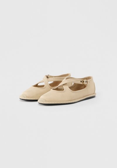 Μπεζ υφασμάτινα μπαλαρίνια με ανοικτές πλευρές, διαθέτουν στρογγυλεμένη μύτη, πλεχτή διακόσμηση από τζούτα στη μύτη και ρυθμιζόμενο λουράκι με πόρπη στο επάνω μέρος του ποδιού.