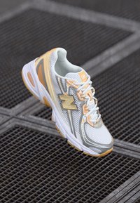 Bela in svetlo oranžna tekaška obuvala New Balance z mrežastim materialom in vezalkami, počivajo na površini iz kovinske rešetke.