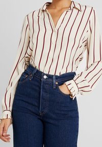Blusa crema con rayas rojas verticales, parte delantera con botones y mangas con puños, combinada con jeans de mezclilla azul oscuro de cintura alta.