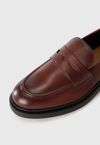 Brown leather loafer with a smooth texture, featuring a decorative strap and stitching details, and a contrasting black rubber sole.