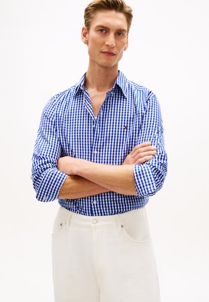 Tânăr cu păr deschis la culoare, purtând o cămașă în carouri albastru și alb și pantaloni albi, stând cu brațele încrucișate pe un fundal alb.