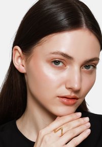Goldener Zickzackring, der an der Hand einer Person mit glattem, dunklem Haar und glatter Haut getragen wird, die ein schwarzes Oberteil vor einem weißen Hintergrund trägt.