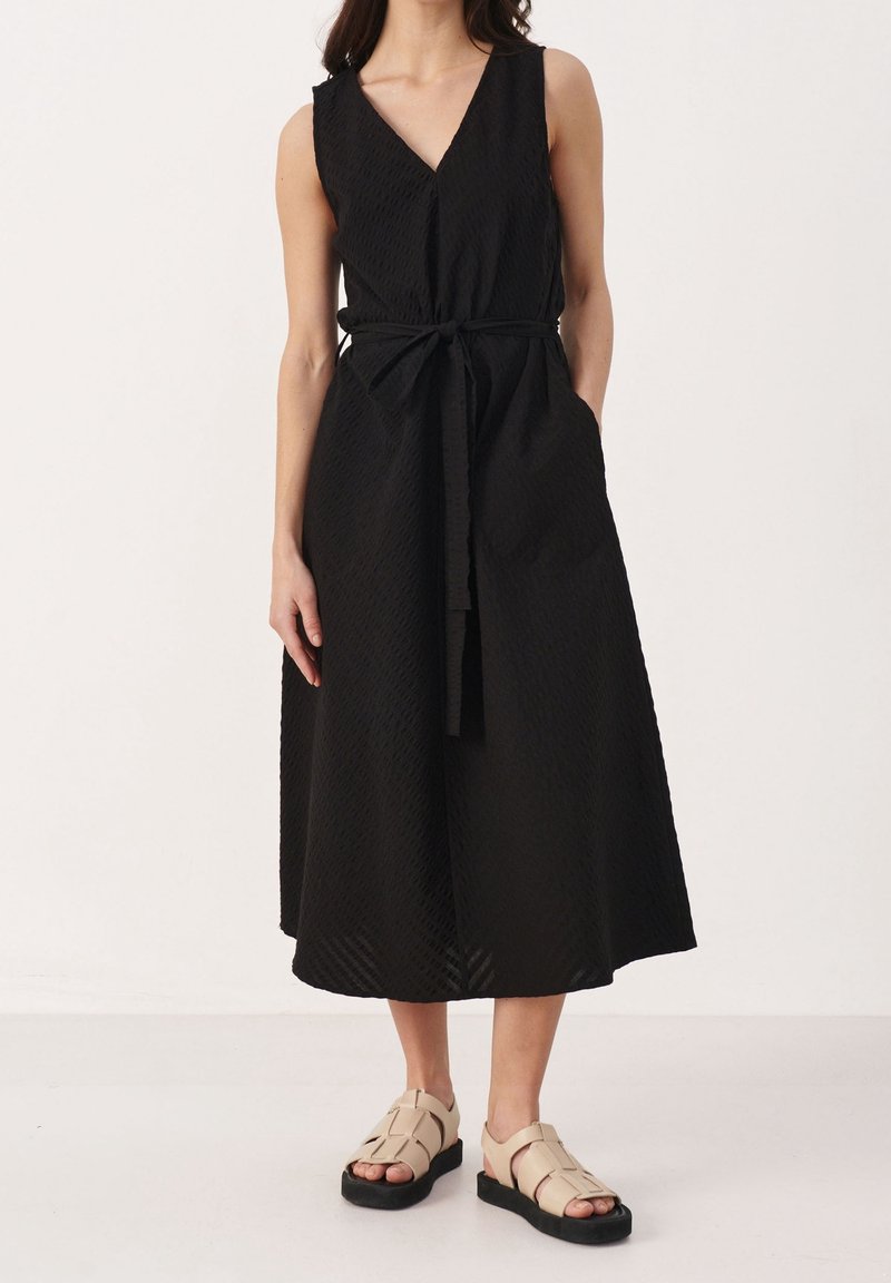 Femme portant une robe midi noire sans manches avec une ceinture à la taille et des sandales ouvertes beige à semelles noires, debout devant un fond uni.