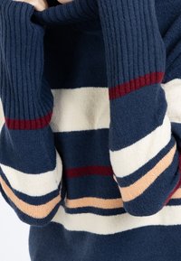 Henkilö, jolla on päällään laivastonsininen villapaita, jossa on leveät valkoiset, punaiset ja persikanväriset vaakaraidat. Kädet osittain pitkien, resorireunaisten hihojen sisällä.