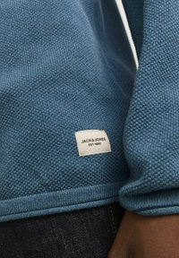 Blå strikket sweater med et tekstureret stof. Har ribbede håndled og kant, samt et hvidt mærke med "JACK & JONES EST. 1990" syet på siden.