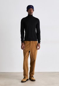 Suéter de cuello alto negro, pantalones de pana marrón y botas beige. Modelo con un gorro de punto azul, de pie sobre un fondo neutro.