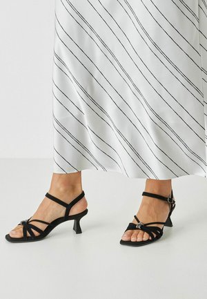 Černé sandály na malém podpatku s pásky, zdobené uzlovým designem a pásky kolem kotníku. Hladká textura s elegantním tvarem.