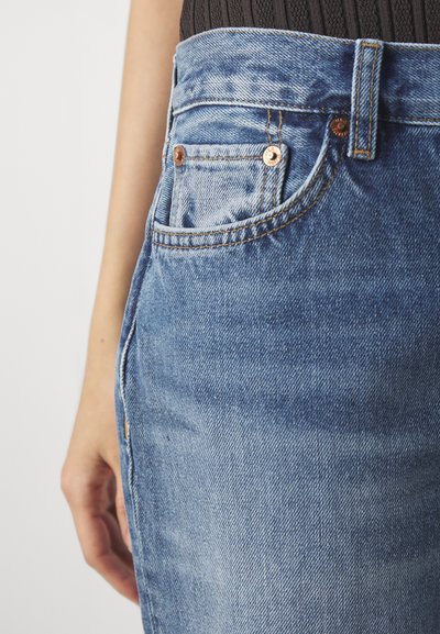 Heledad sinine denim, mille lähedane vaade, mis sisaldab kontrastset oranži õmblust, väikest taskut ja vasktooni riistvara detaile.