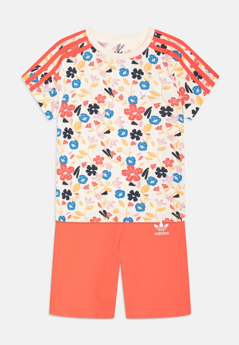 Top a maniche corte con stampa floreale in crema con fiori rossi e blu, abbinato a pantaloni corti corallo con logo Adidas bianco e righe di branding.