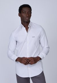 Chemise blanche à boutons avec un motif discret de pois bleus, col standard et manches longues. Associée à un pantalon gris.