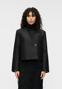 Μαύρο κροπ σακάκι με υπερμεγέθη μανίκια, που διαθέτει ένα μόνο κυκλικό κουμπί, φορεμένο πάνω από μια υφασμάτινη μαύρη φούστα με κρόσσια.