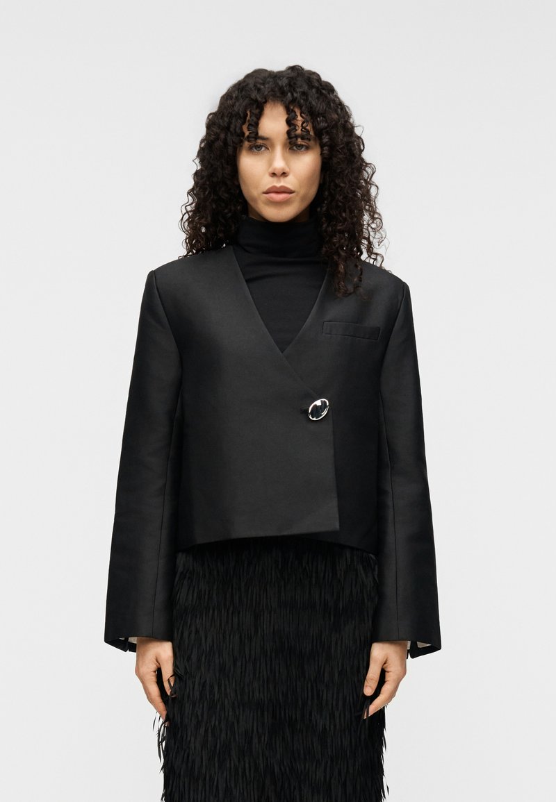 Μαύρο κροπ σακάκι με υπερμεγέθη μανίκια, που διαθέτει ένα μόνο κυκλικό κουμπί, φορεμένο πάνω από μια υφασμάτινη μαύρη φούστα με κρόσσια.