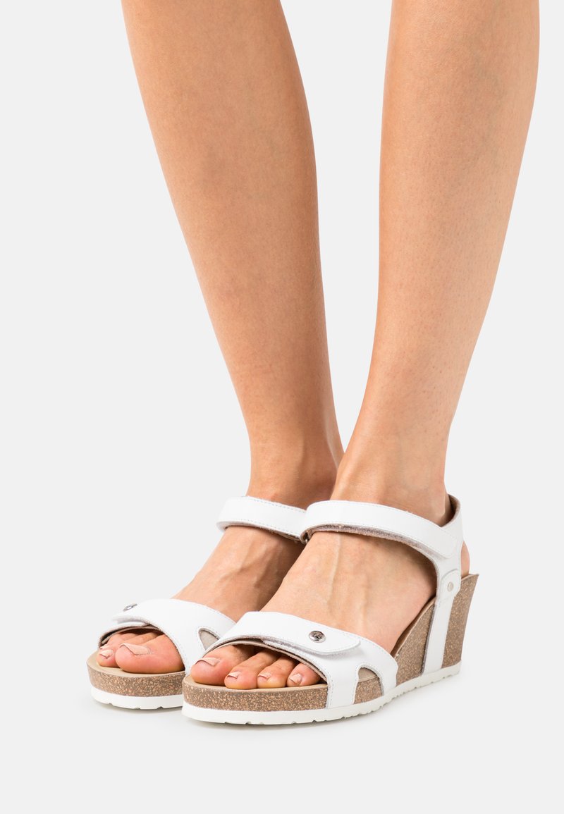 Sandalias de cuña blancas con correas de cuero, diseño de punta abierta y suela de aspecto de corcho. Cuenta con correas ajustables en los tobillos y acentos decorativos de plata.