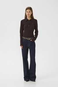 Young woman standing with hand in pocket wearing a dark brown long-sleeve polo shirt, blue wide-leg jeans, and a brown belt.