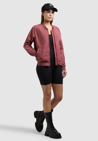 Woman standing wearing a maroon bomber jacket, black cycling shorts, black combat boots, and a black baseball cap on a light background.