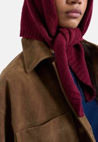 Femme portant une écharpe tricotée bordeaux nouée sous le menton et une veste en daim marron sur un haut bleu.