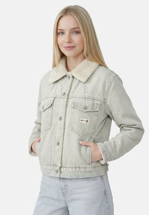 Ung kvinde med langt blond hår iført en lysegrå denimjakke med hvid fleecekrave og hænderne i lommerne.