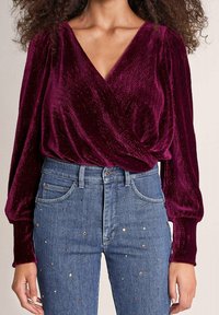 Bordeaux fluwelen wikkelblouse met lange mouwen en een diepe V-hals, gecombineerd met lichtblauwe jeans met meerkleurige edelsteenversieringen.