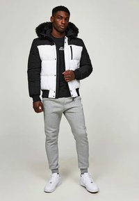Men's puffer jacket in black and white, featuring a faux fur-lined hood, front pockets, and quilted texture. Paired with gray sweatpants.