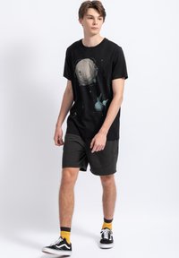 Zwarte katoenen T-shirt met een grafisch ontwerp van een planeet en sterren. Gecombineerd met donkergrijze shorts, geel-zwarte sokken en zwarte sneakers.