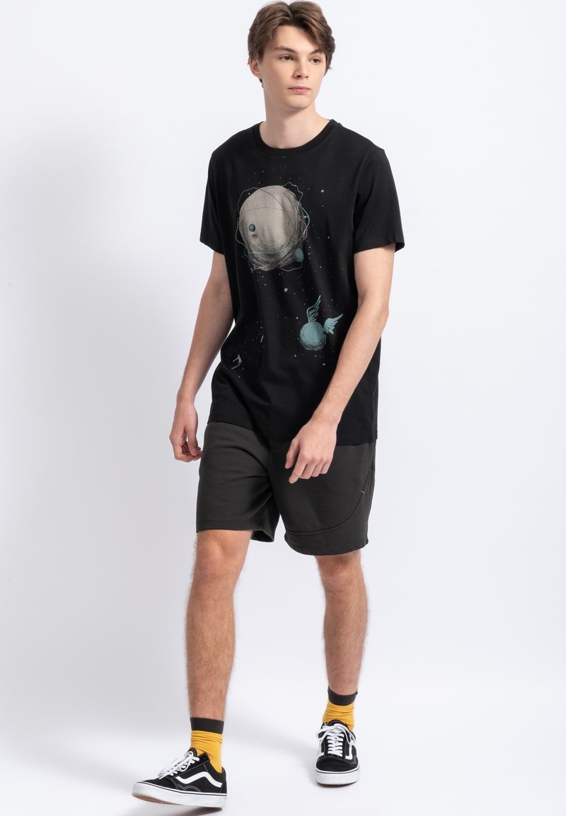 Zwarte katoenen T-shirt met een grafisch ontwerp van een planeet en sterren. Gecombineerd met donkergrijze shorts, geel-zwarte sokken en zwarte sneakers.