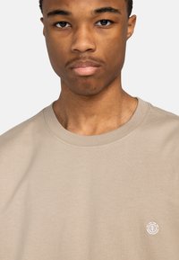 Beige Baumwoll-T-Shirt mit Rundhalsausschnitt und dezentem weißen Logo auf der linken Brust. Angenehme Textur mit kurzen Ärmeln.
