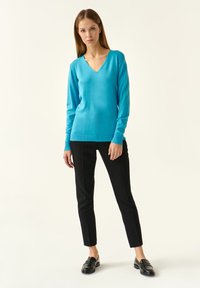 Pulover turcoaz cu decolteu în V, realizat dintr-un material moale, cu mâneci lungi, purtat cu pantaloni negri ajustați și mocasini negri. Design minimalist.