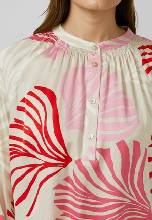 Woman wearing a cream blouse with large red and pink abstract leaf patterns and a round neckline with button closure.