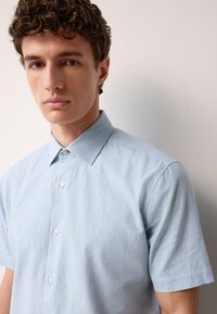 Camicia a maniche corte, azzurro chiaro con righe verticali bianche, dotata di collo classico e texture liscia.