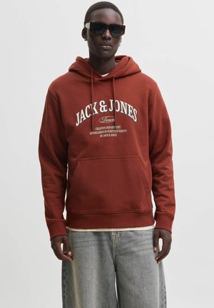 Personne portant des lunettes de soleil foncées, un sweat à capuche rouille Jack & Jones avec un texte blanc et un jean bleu clair sur un fond uni.