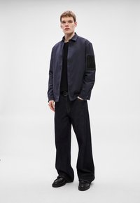 Lahka mornarsko modra jakna s stoječim ovratnikom, kombinirana s črno srajco in ohlapnimi temnimi džins hlačami. Črne copate brez vezalk dopolnjujejo videz.