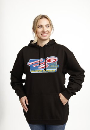 Uśmiechnięta kobieta, ubrana w czarną bluzę z kapturem z czerwono-niebieską grafiką statku kosmicznego i napisem "NCC-1701 Warp Speed", ręce w kieszeniach na białym tle.