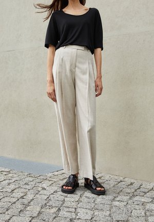Mujer con pantalones beige sueltos plisados, camiseta negra de manga corta y sandalias negras gruesas, de pie sobre pavimento de adoquines.