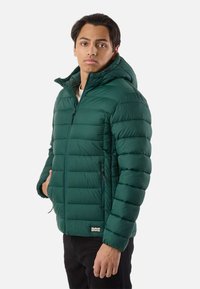 Jeune homme portant une veste matelassée à capuche vert foncé avec des fermetures éclair, debout devant un fond clair uni, légèrement de profil.