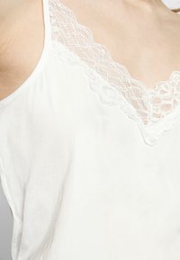 Witte camisole met een kanten rand bij de hals. De stof is zacht met een lichte glans en de schuine bandjes zijn dun, wat een delicate uitstraling geeft.