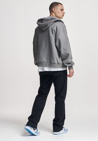 Man wearing grey hoodie, white T-shirt, black pants, and white and blue sneakers standing with back turned and head in profile.