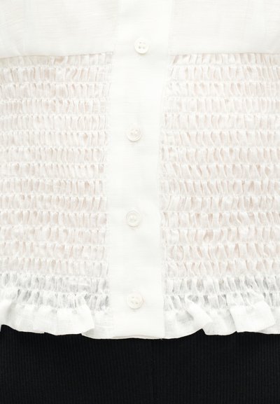 Chemise blanche boutonnée avec des panneaux en tissu froncé texturé et un ourlet à volants, portée sur un pantalon noir côtelé.