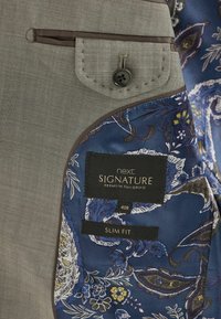 Interior de blazer de corte ajustado con forro floral azul, exterior gris con acabado texturizado, detalle de bolsillo y etiquetas de marca.