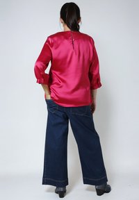 Woman with dark hair in ponytail wearing a shiny magenta blouse with buttons on the back and dark wide-leg jeans with black heeled boots.