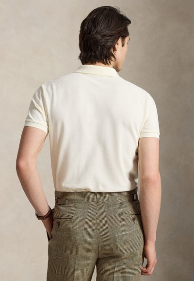 Mężczyzna o ciemnych włosach, ubrany w kremową koszulkę polo z krótkim rękawem włożoną w brązowe, kratkowane spodnie, stoi z rękami w kieszeniach.