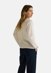 Femme avec de longs cheveux portant un pull en tricot beige ample et un pantalon large sombre, se tenant avec sa main dans sa poche, regardant sur le côté.