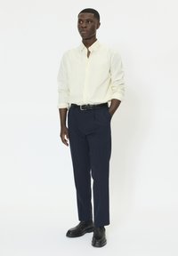 Chemise pâle jaune à col, pantalon bleu marine foncé à rayures fines, ceinture tressée noire, chaussures noires, pose debout avec les mains dans les poches, fond neutre.