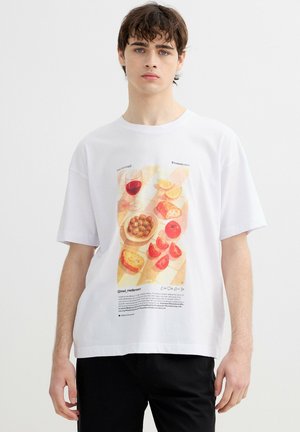 Joven con una camiseta blanca con un estampado de comida mediterránea que muestra pan, aceitunas, rodajas de tomate, gajos de limón y vino tinto.