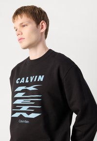 Sweatshirt preto com gola redonda, apresentando tipografia azul em destaque e padrões gráficos. Feito de material suave com uma textura lisa.