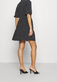 Robe noire à manches courtes avec imprimé de cœurs blancs, taille cintrée, jupe évasée. Le mannequin porte des talons noirs ouverts, mettant en valeur l'ourlet de la robe.