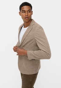 Beige Cordhosenblazer mit Reverskragen, einer Brusttasche und Zwei-Knopf-Verschluss, kombiniert mit einem weißen T-Shirt und olivgrünen Hosen.