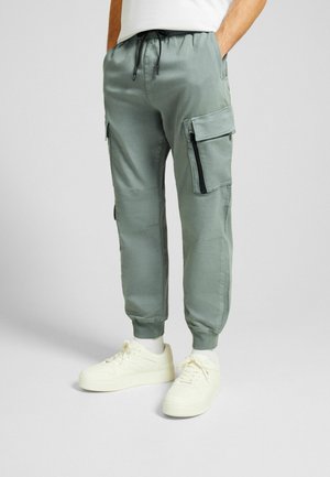 Pantalones cargo verde claro con bolsillos con cremallera, cinturilla elástica y puños. Combinados con zapatillas blancas. Textura suave y diseño ajustado.